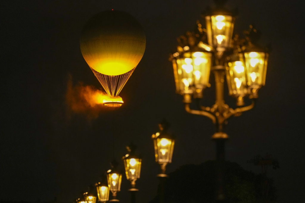 Color photograph aimed upwards from the ground on a dark night. What is visible is a large inflated balloon tethered by many strings to a basket holding light and giving off steam which lights the balloon from below with a lovely glow. A row of old-style streetlamps are in a line from the top left to the bottom right lit up to match the balloon. 